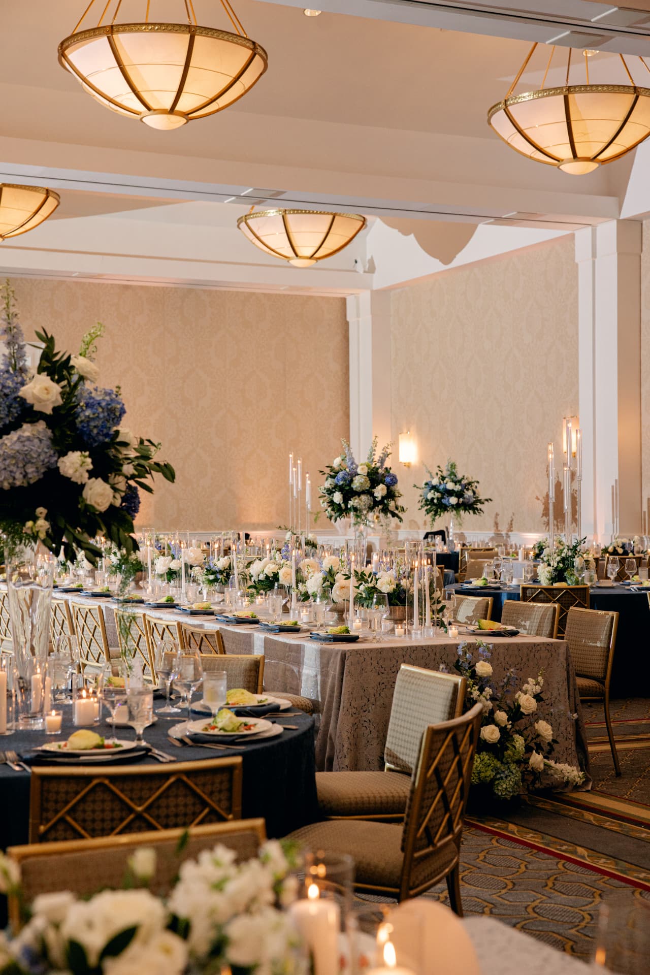 Candlelit reception tables with blue and white florals beneath gold chandeliers at Chuck and John's wedding